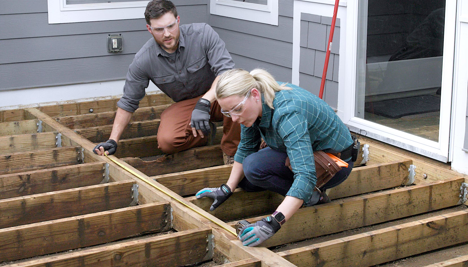 Two workers wearing safety glasses and gloves measuring a wooden deck frame during outdoor construction.