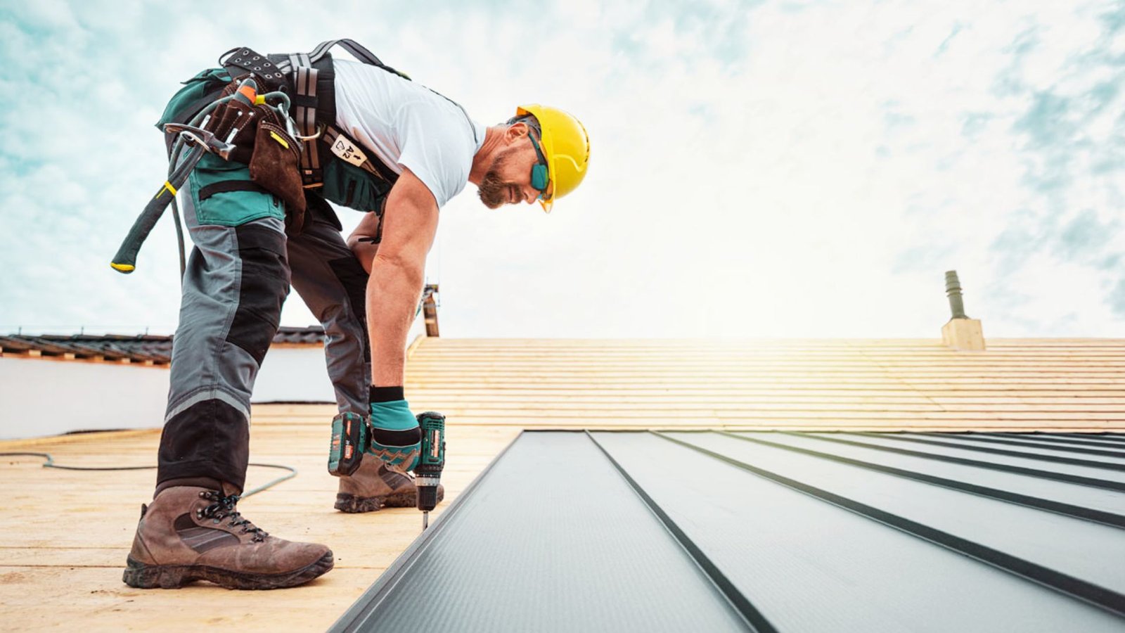 Construction worker installing standing seam metal roofing panels on a residential roof using a power drill while wearing safety gear and a tool harness