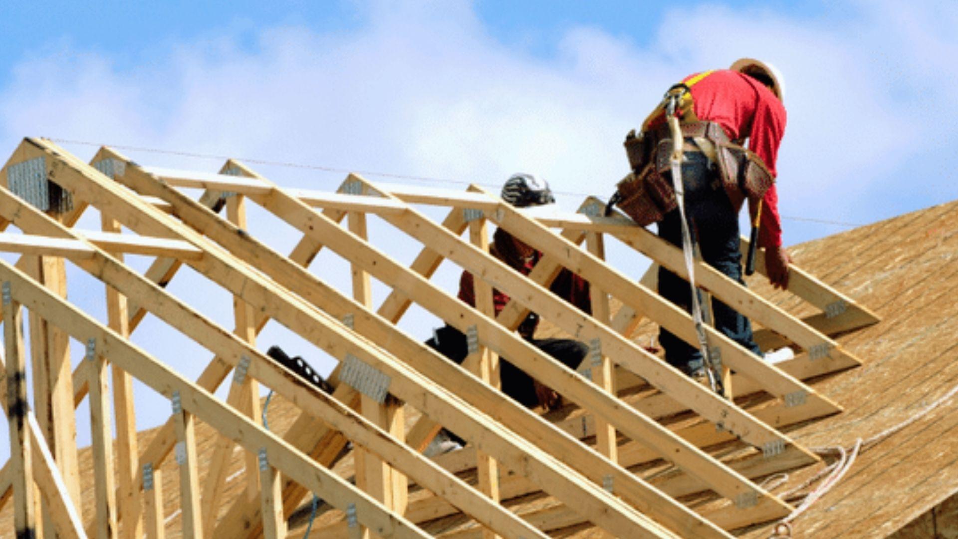Roofers installing wooden roof trusses on house construction wearing safety harnesses under blue sky