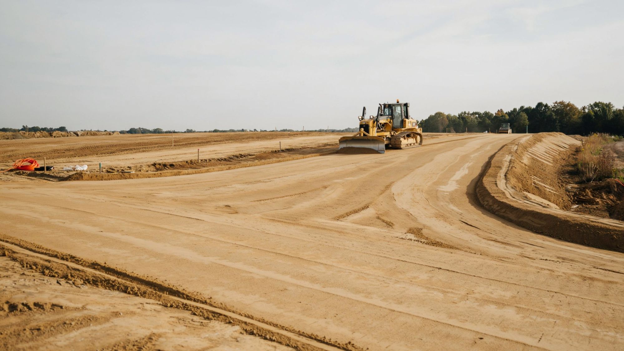 Tracked excavator digging trench in dusty terrain with embankments and construction fencing.