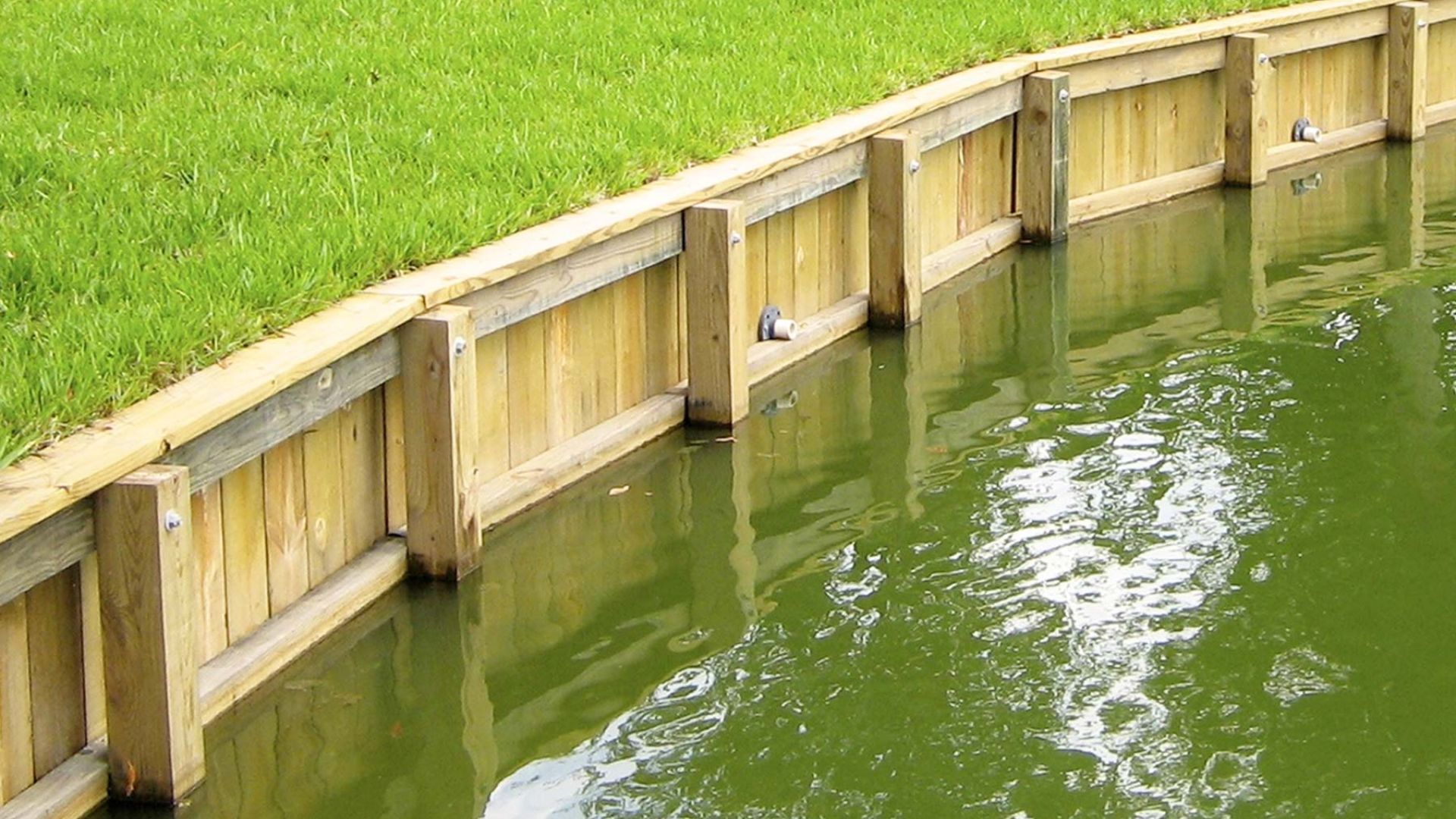 Wooden bulkhead seawall retaining green grass lawn above murky green canal water.