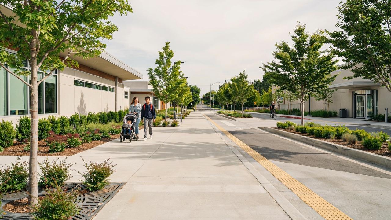 Two people pushing strollers on a wide modern walkway lined with young trees and landscaped buildings.