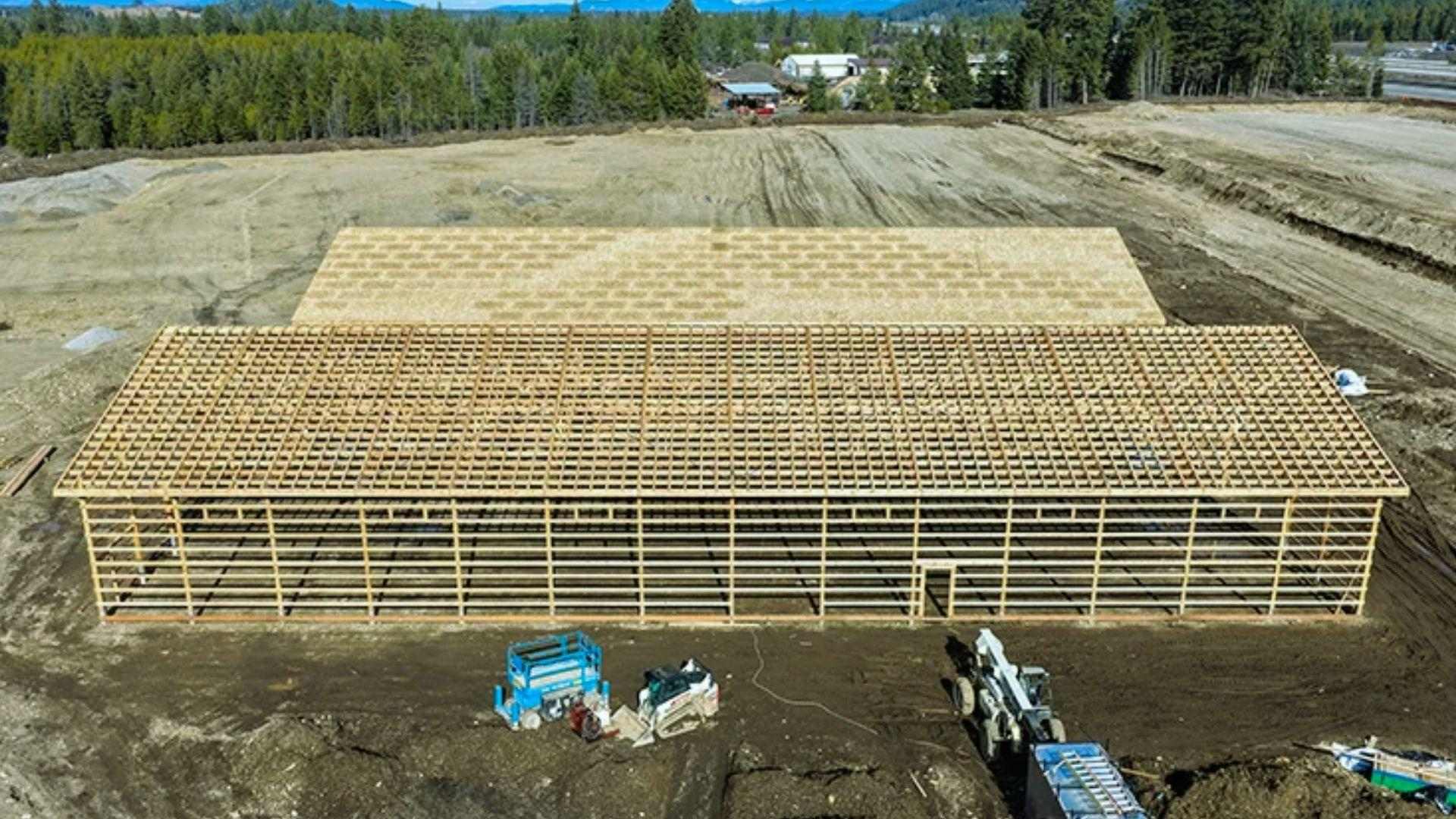 Aerial view of large wooden pole barn construction with roof purlins, machinery on dirt site with mountains background