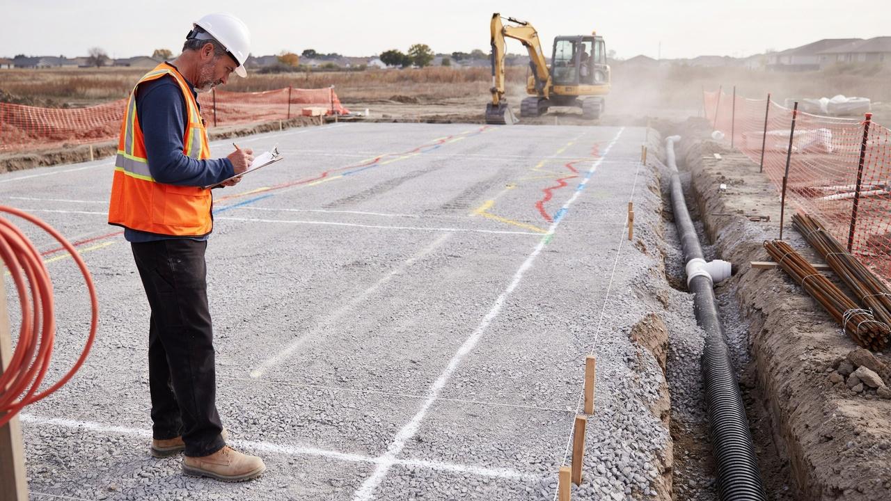 Construction worker in safety vest inspecting gravel foundation site with excavator, pipe trench and rebar