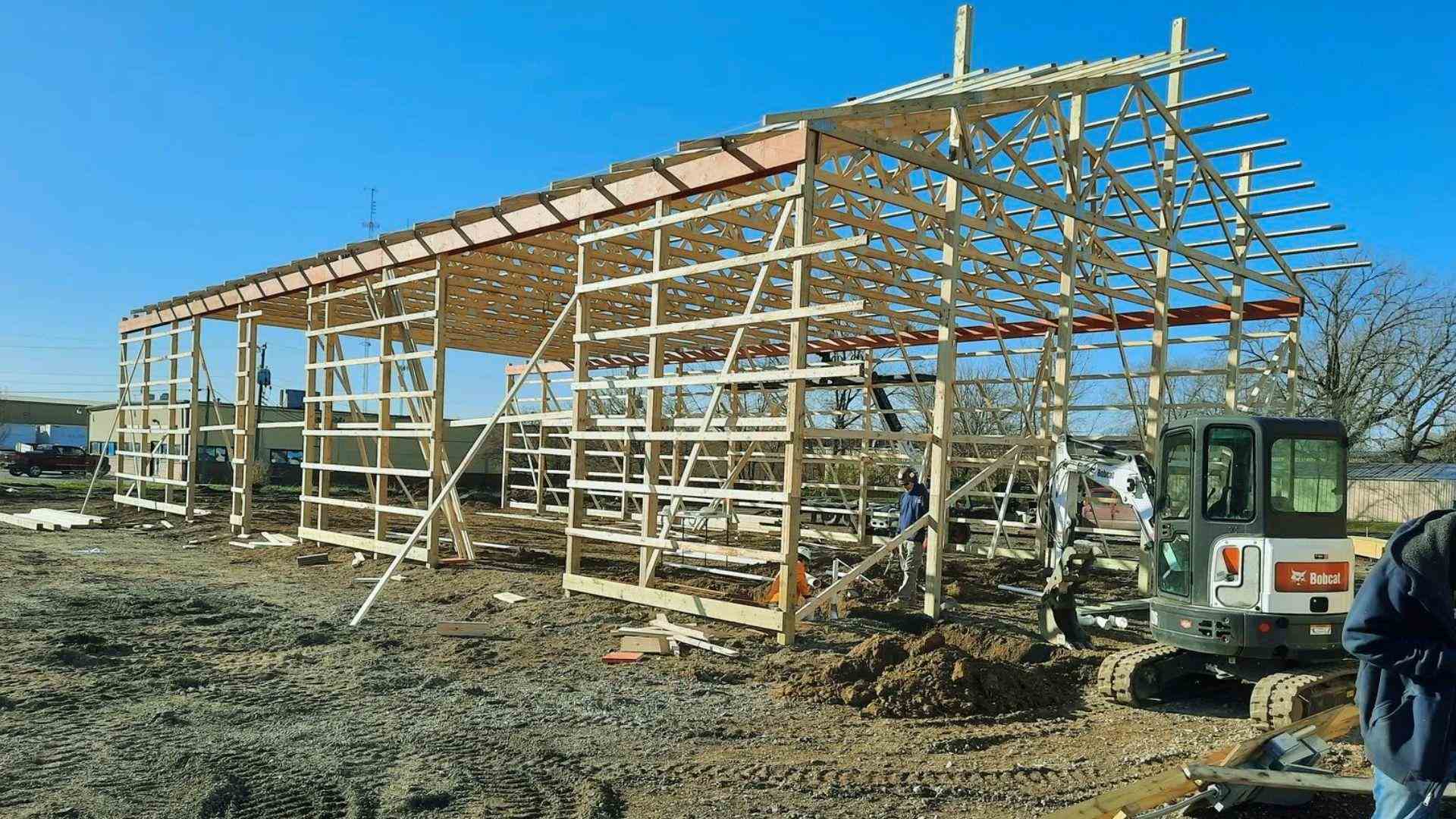 Wooden pole barn frame construction with roof trusses and excavator on dirt job site under blue sky