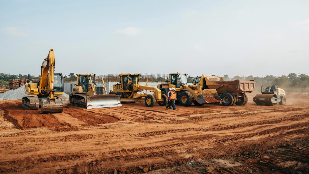 Lineup of heavy construction equipment on red dirt site with workers inspecting excavator, bulldozer, grader, loader, dump truck and roller.