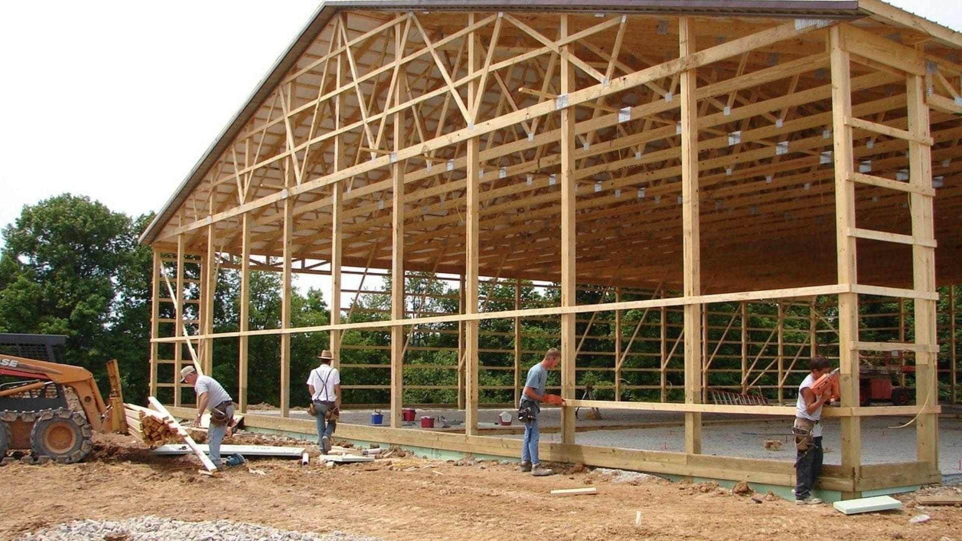 Construction workers building wooden pole barn frame with trusses on dirt job site