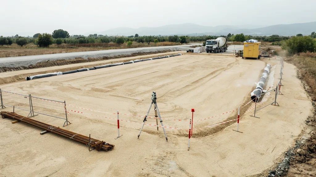 Survey tripod on sandy lot with rebar bundles, drainage pipes, concrete mixer truck in distance.