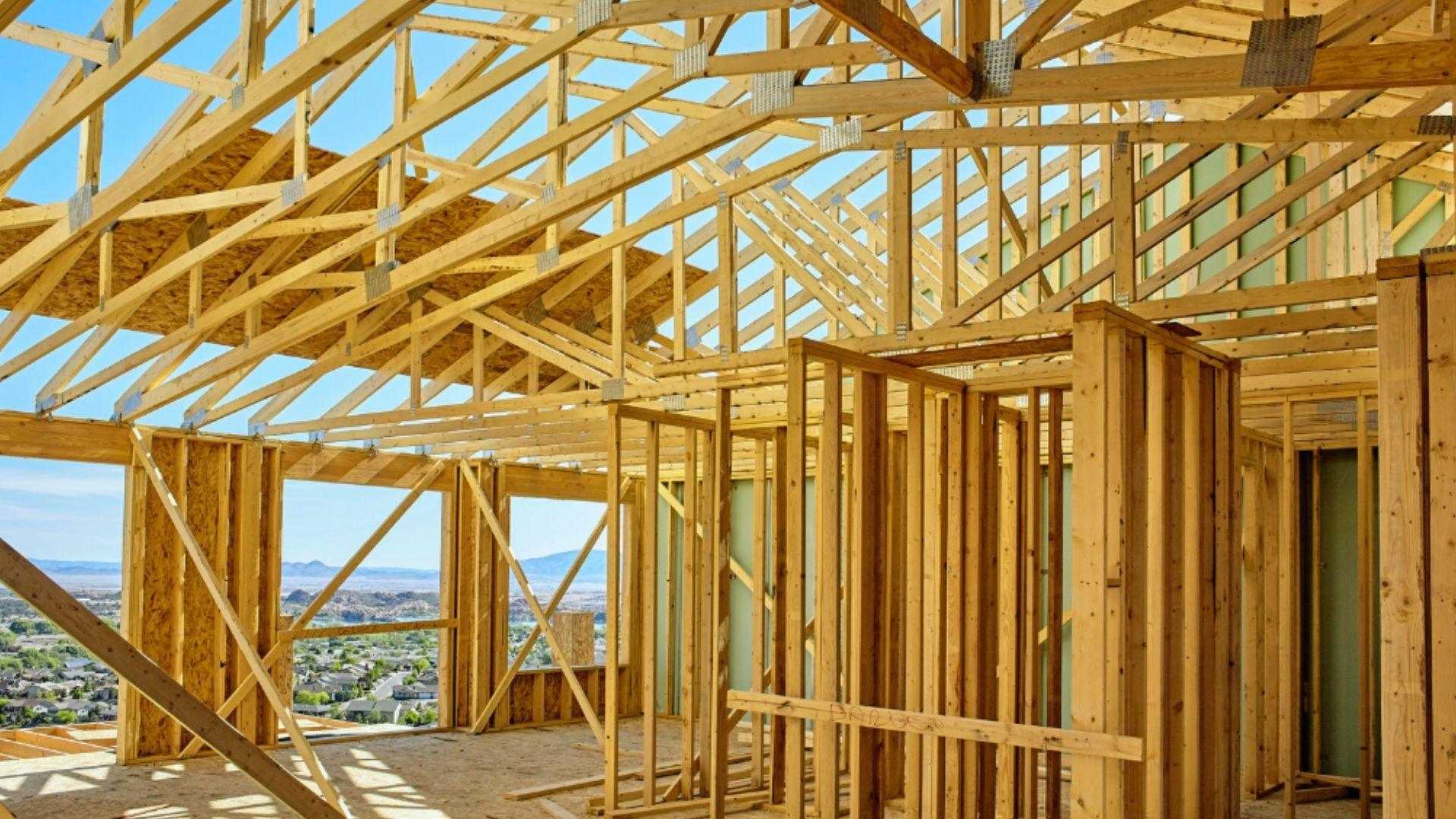 Wooden house framing with roof trusses and wall studs overlooking suburban landscape under blue sky