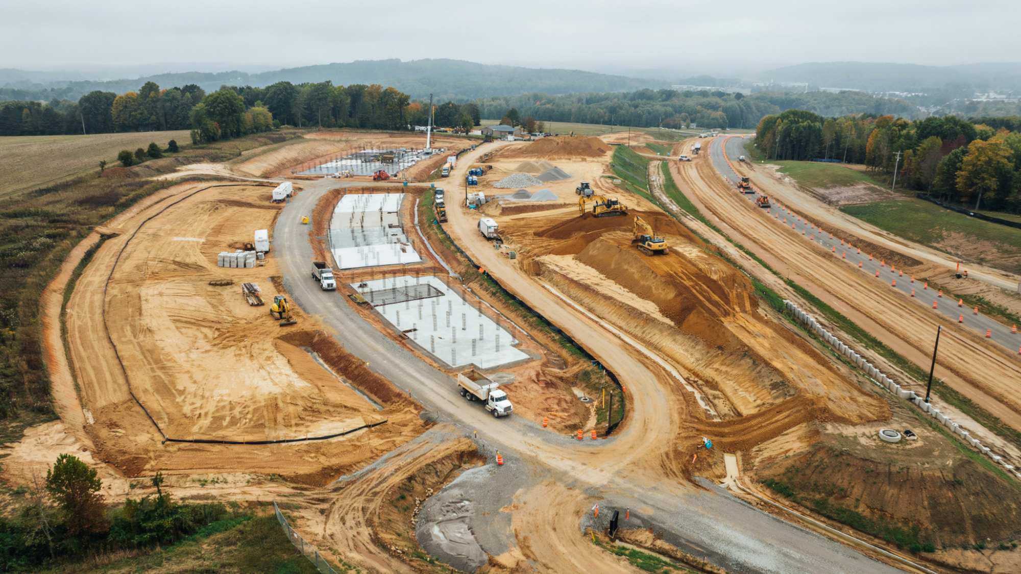 Aerial view of expansive highway construction site with concrete foundations, excavators, dump trucks, and earthworks.
