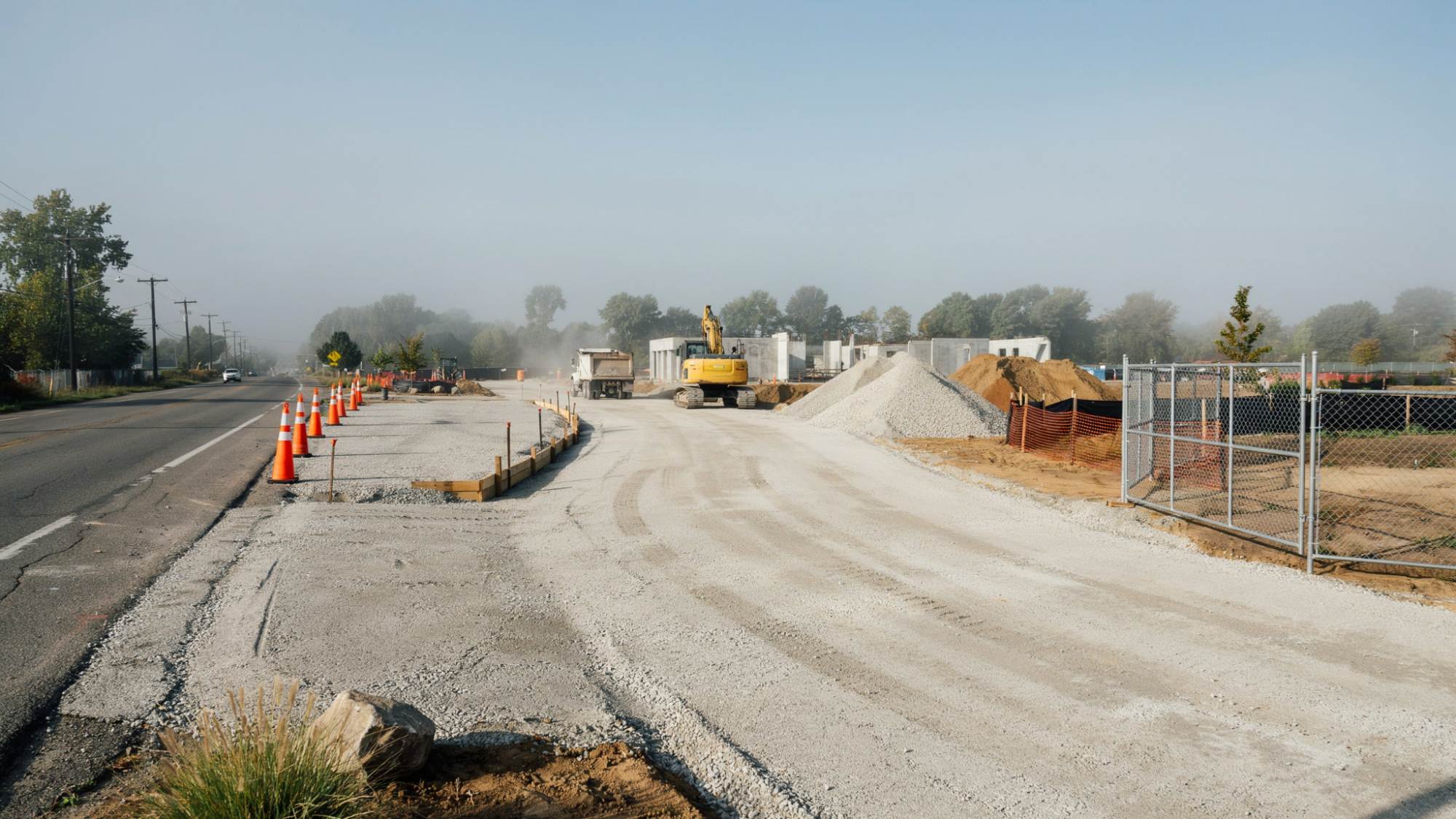 Excavator and dump truck on foggy roadside construction site with gravel piles, cones, and fencing.