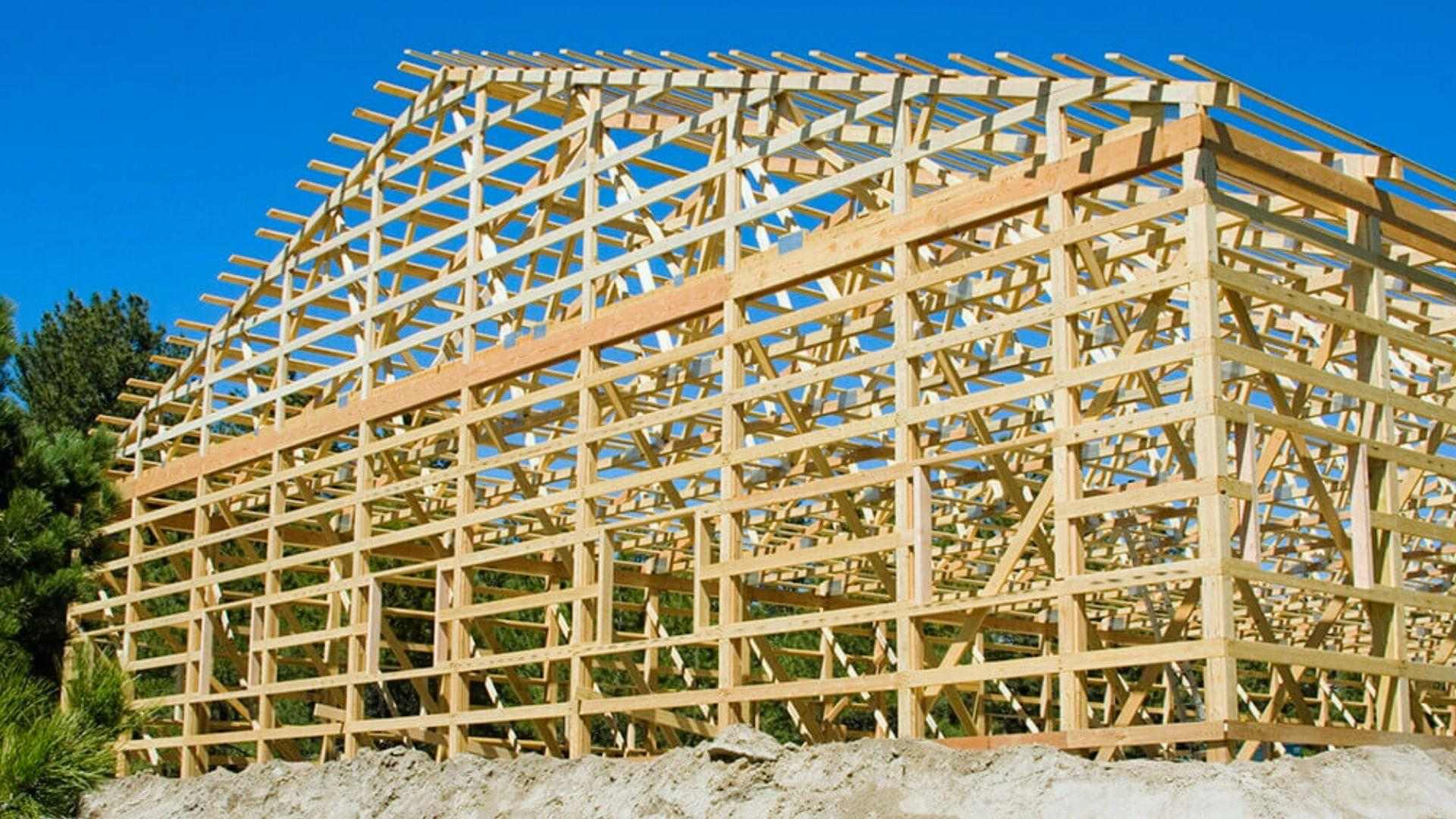 Wooden pole barn frame construction with roof trusses and purlins on dirt mound under blue sky