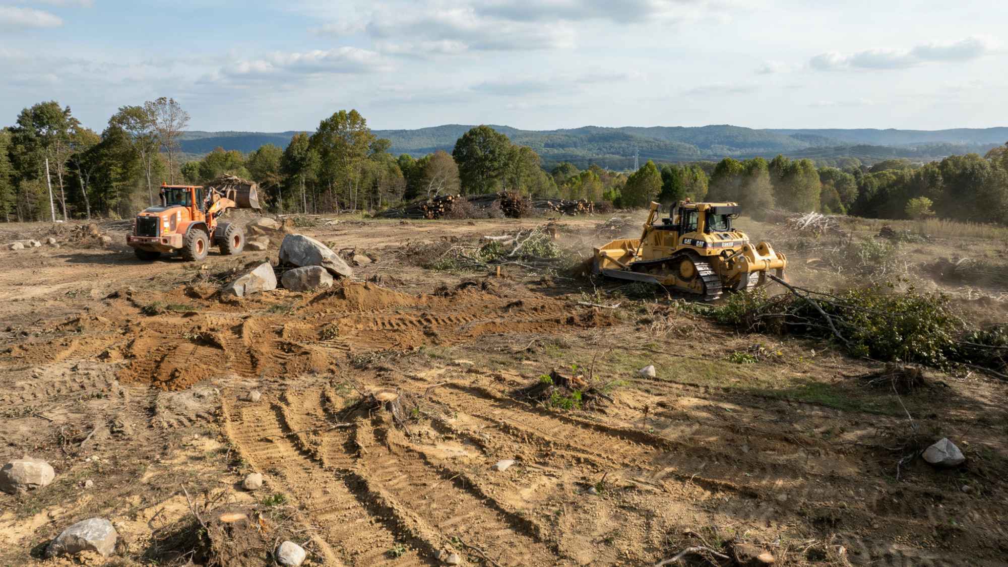 Orange wheel loader and yellow bulldozer clearing brush and rocks on dusty deforested construction site.