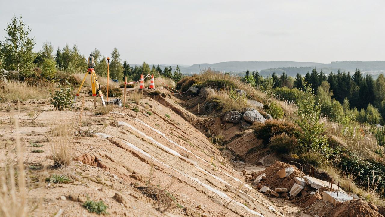 Surveyor tripod on eroded hillside with orange cones, construction debris, forested valley in distance.