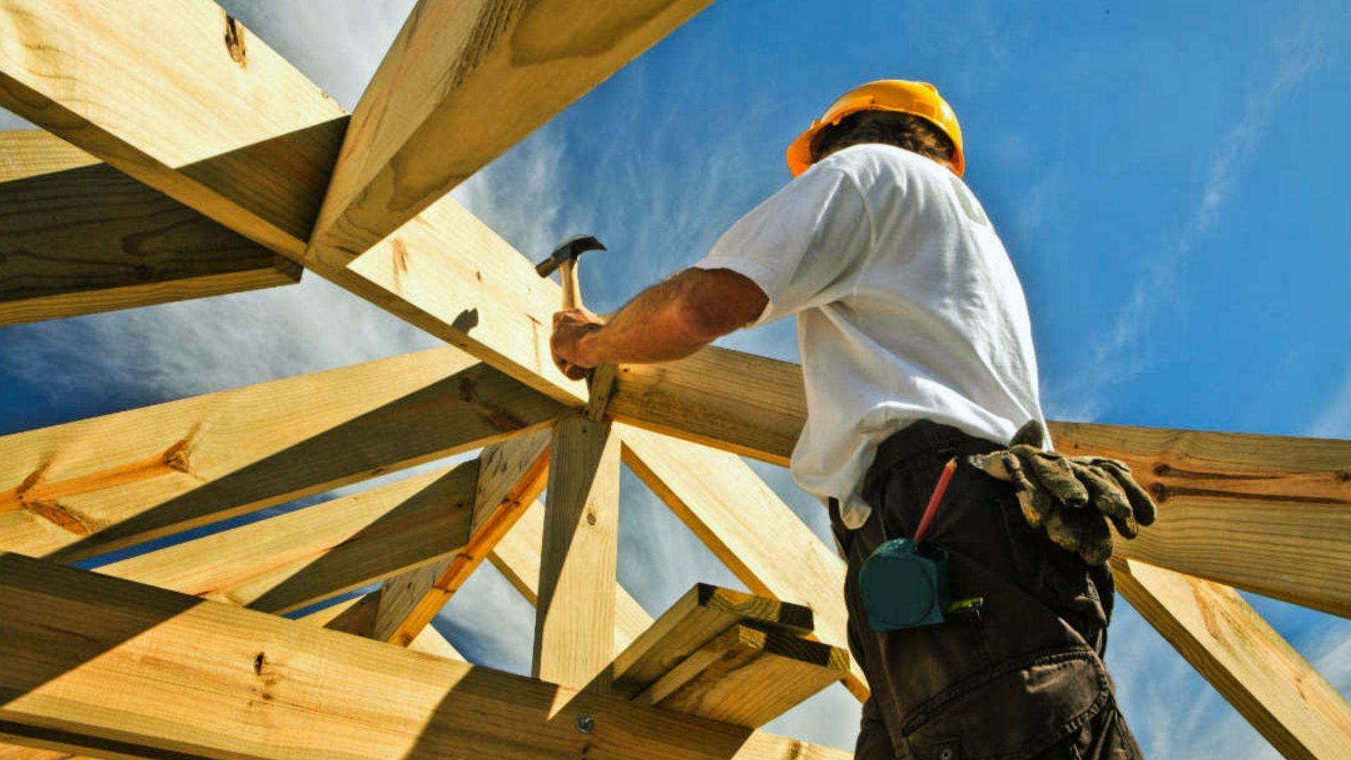 Construction worker hammering wooden roof trusses wearing yellow hard hat under blue sky