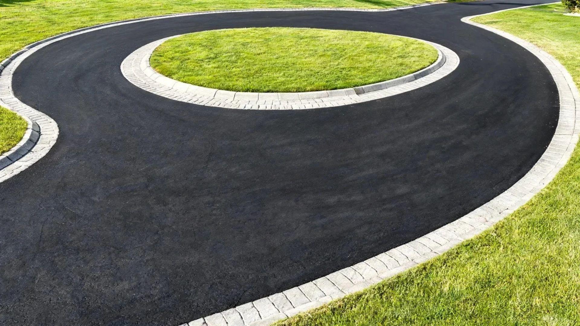Dark asphalt horseshoe driveway with a contrasting paver border beside a green lawn.
