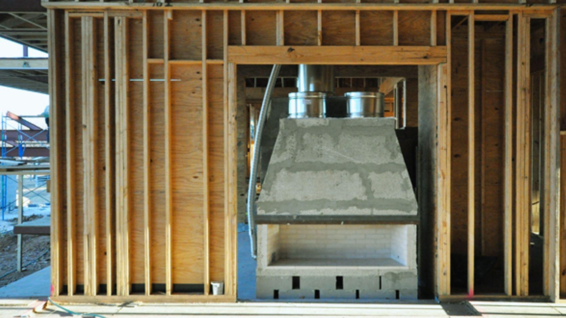 Concrete fireplace under construction inside wooden wall framing with metal ductwork and scaffolding visible