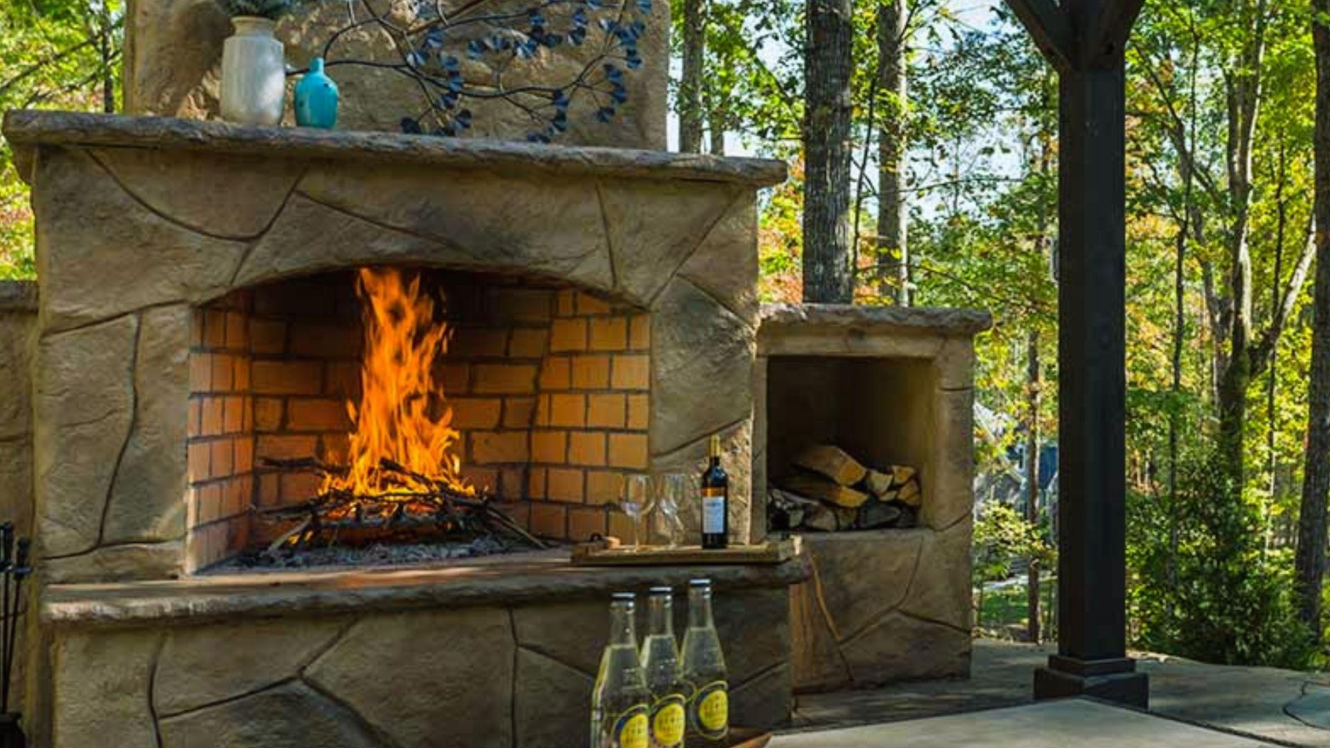 Outdoor stone fireplace with roaring fire, built-in wood storage, and drinks on patio surrounded by forest