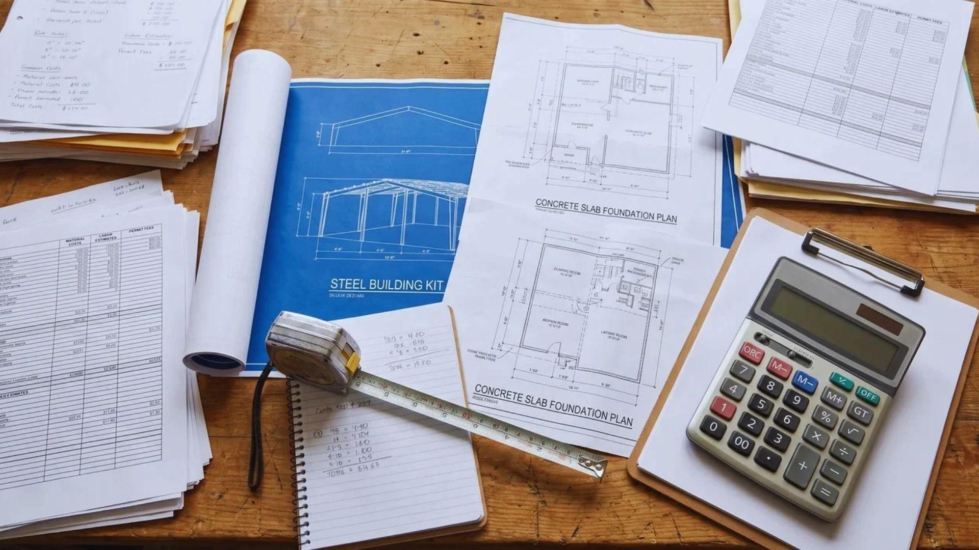 Building plans, a calculator, a measuring tape, and cost-estimate documents arranged on a work surface.